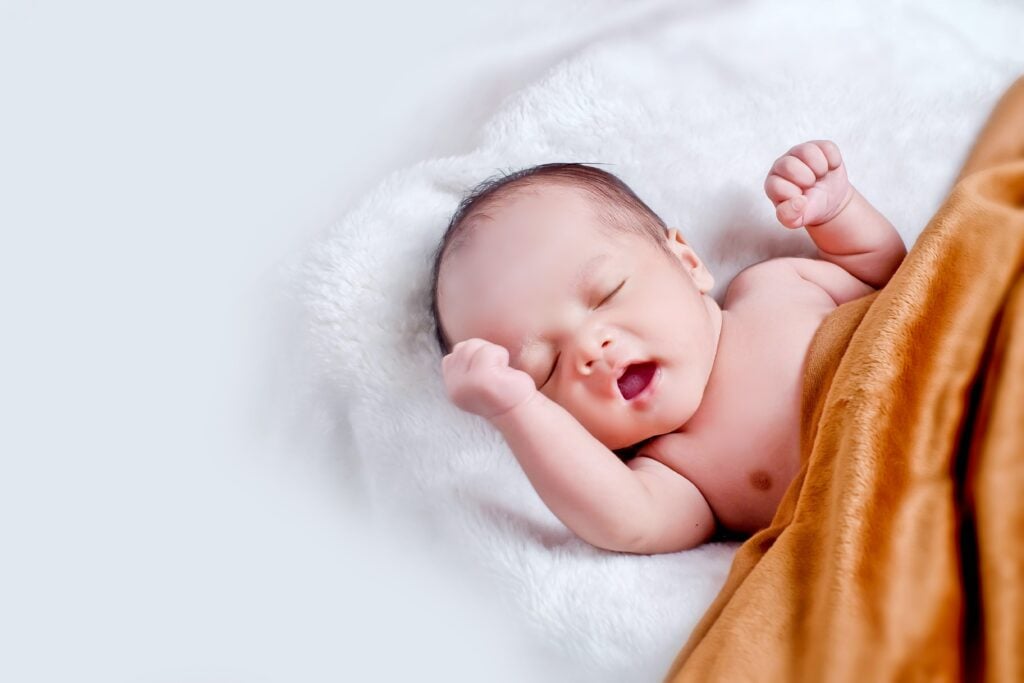 Baby laying on a plush white blanket, yawning with fist in the air. By 4 months old, a baby is looking for structure in their daily routine. This is the optimal time to start sleep training with an in-home baby sleep expert.