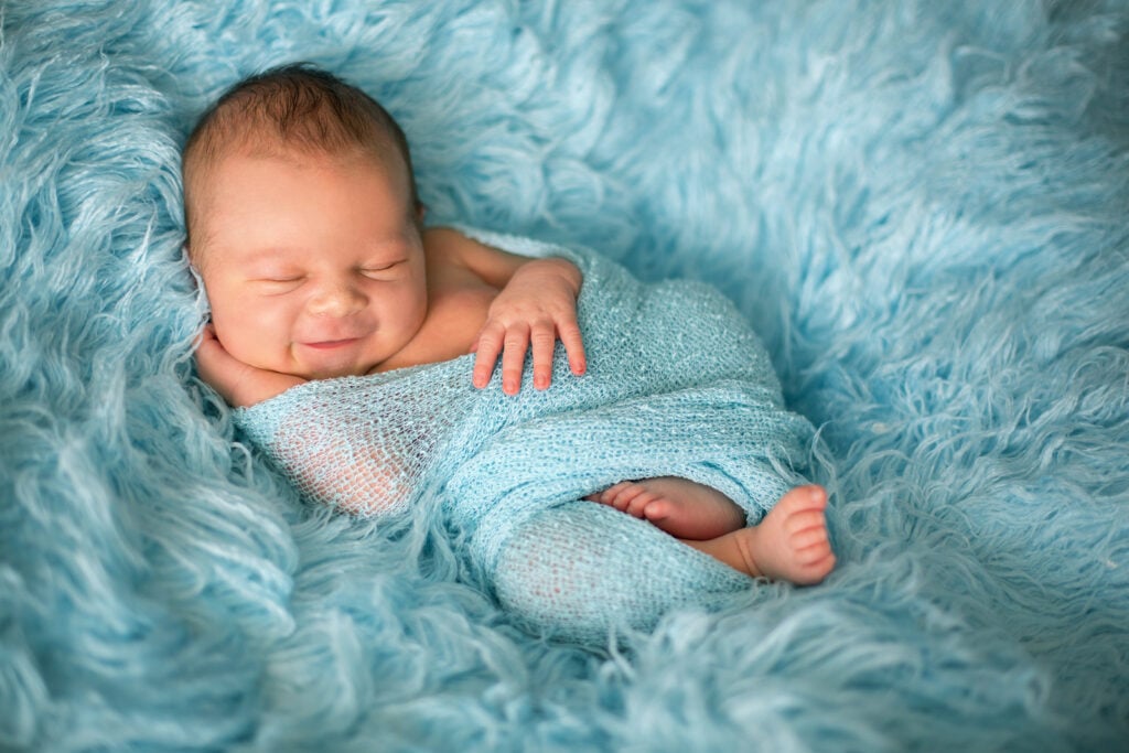 Happy smiling newborn baby in wrap sleeping. Swaddling can help with nighttime sleep training and naps. The Peaceful Sleeper can help with your sleep problems for infants, toddlers and adults.