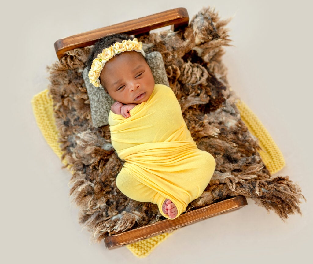 Newborn baby swaddled in yellow lying in cradle. She sleeps well after being sleep trained as a newborn by The Peaceful Sleeper.