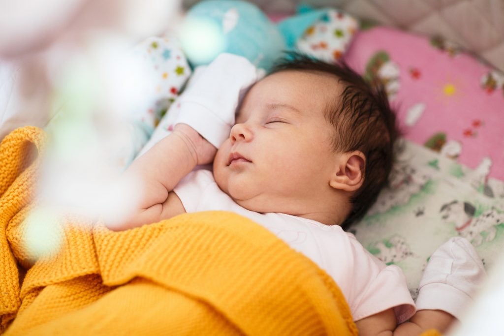 Newborn baby sleeping on her back, swaddled in yellow blanket. She learned to self-soothe using sleep training. Learn more about no cry sleep training in this blog post by The Peaceful Sleeper. 